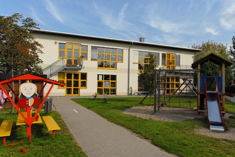 Das Gebäude von der Kita Kleine Strolche und der dazugehörige Spielplatz, mit einem Klettergerüst und einer eingebauten Rutsche. 